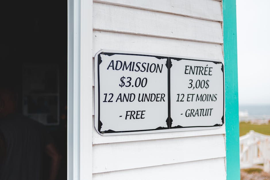 Close-up of a bilingual sign attached to the white wooden exterior of a building, displaying flower arrangements pricing. The sign reads 'ADMISSION $3.00 12 AND UNDER - FREE' in English and 'ENTRÉE 3