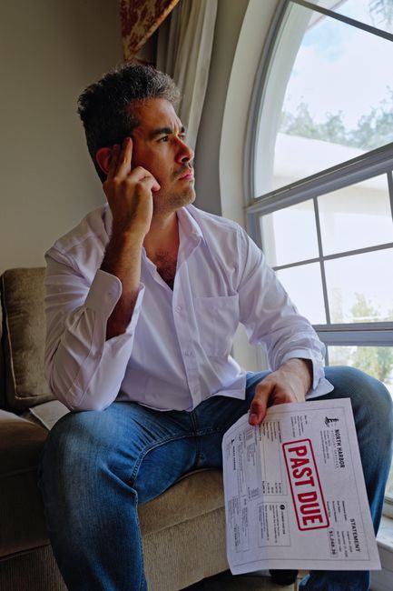 A man with dark, curly hair and a serious expression sits on a beige sofa, resting his right hand on his temple while holding a document in his left hand. He is dressed in a white button-up shirt and 
