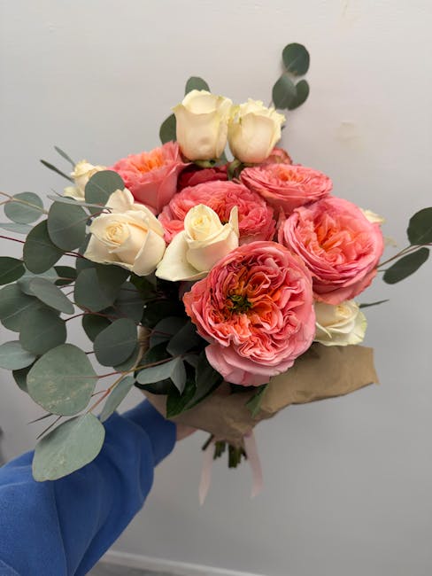 A hand in a blue sleeve holds a bouquet of fresh flowers wrapped in brown paper, featuring large pink ranunculus blossoms with ruffled petals, creamy white roses with tightly closed buds, and eucalypt