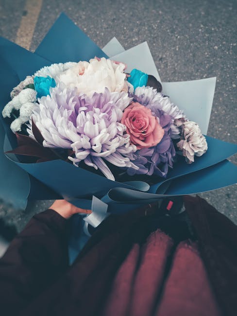 A vibrant floral bouquet featuring large, soft lavender chrysanthemums, a delicate pink rose, and smaller light gray carnations, accented with bright blue hyacinths. The flowers are arranged in a lush