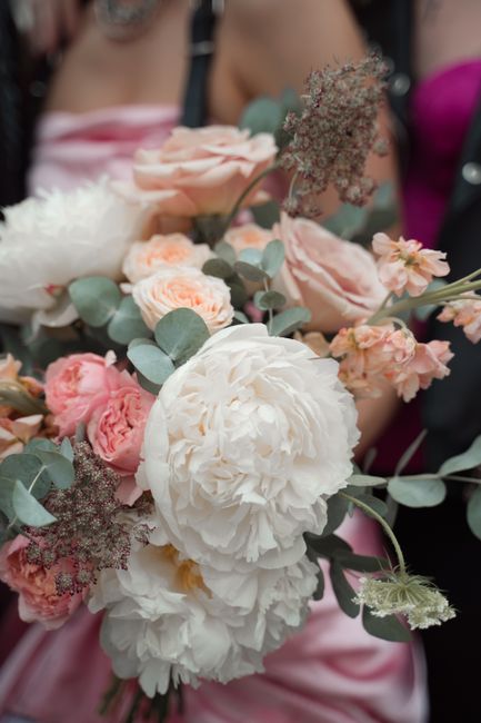 A close-up of a floral bouquet featuring large, soft white peonies with ruffled petals, complemented by blush pink roses and smaller peach-toned flowers. The arrangement includes sprigs of eucalyptus