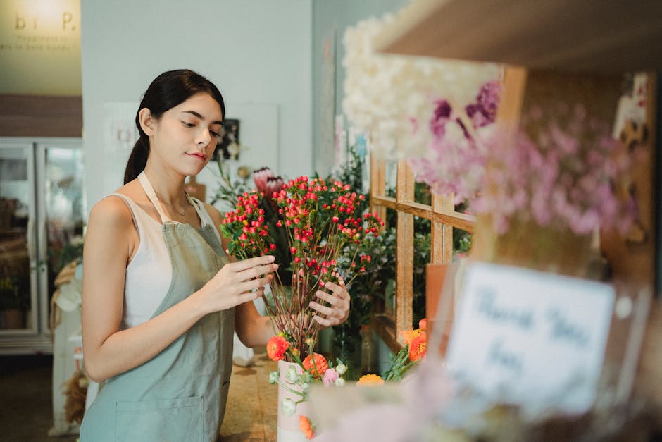 A young woman with dark hair, dressed in a white sleeveless top and a light green apron, is arranging a bouquet of small pink and red flowers with green stems. She is standing behind a wooden counter