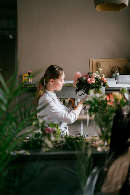 A woman with light brown hair tied back, wearing a white shirt, is holding a large, round bouquet of fresh pink and cream roses, along with greenery, inside a well-lit florist studio. The bouquet feat